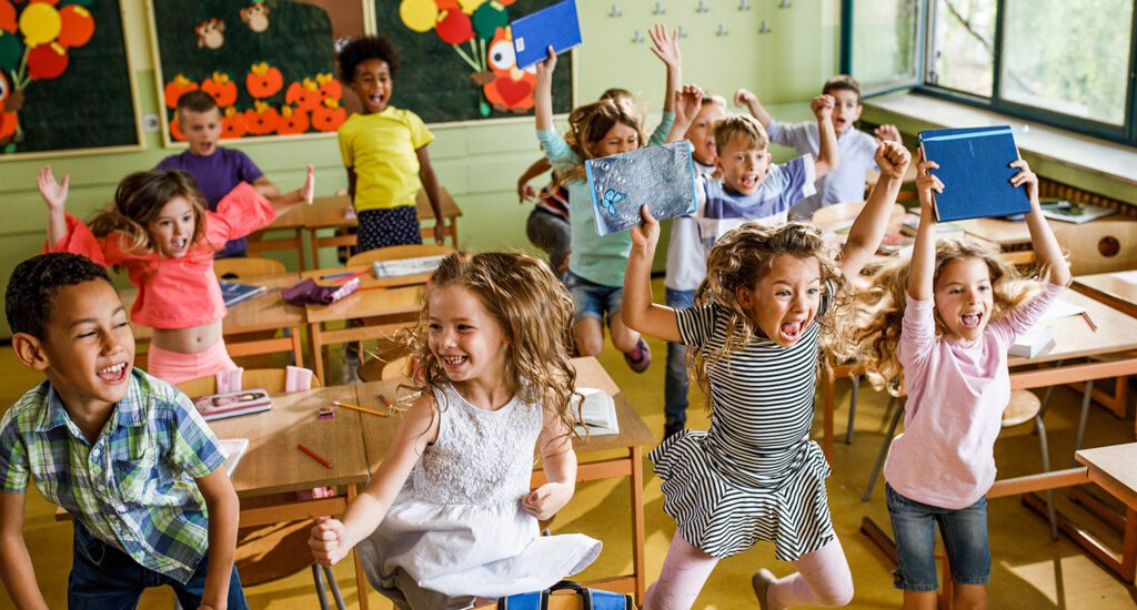 Large group of cheerful school children having fun while jumping among desks in the classroom.